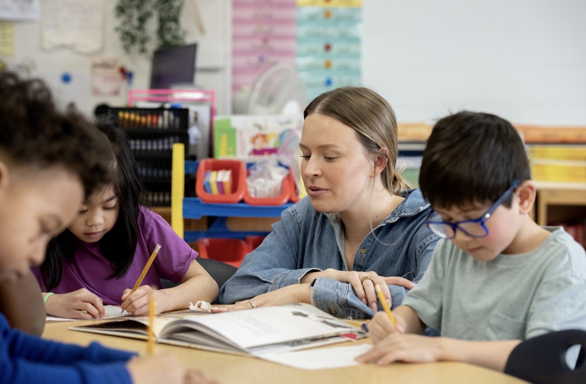 A teacher works closely with four young children writing in their workbooks at a classroom table