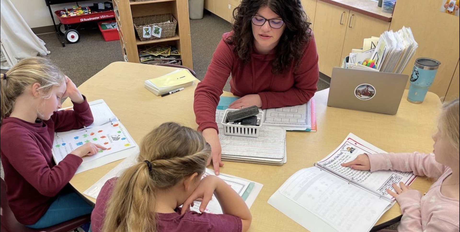 A teacher leads a small group of three children working in their reading workbooks