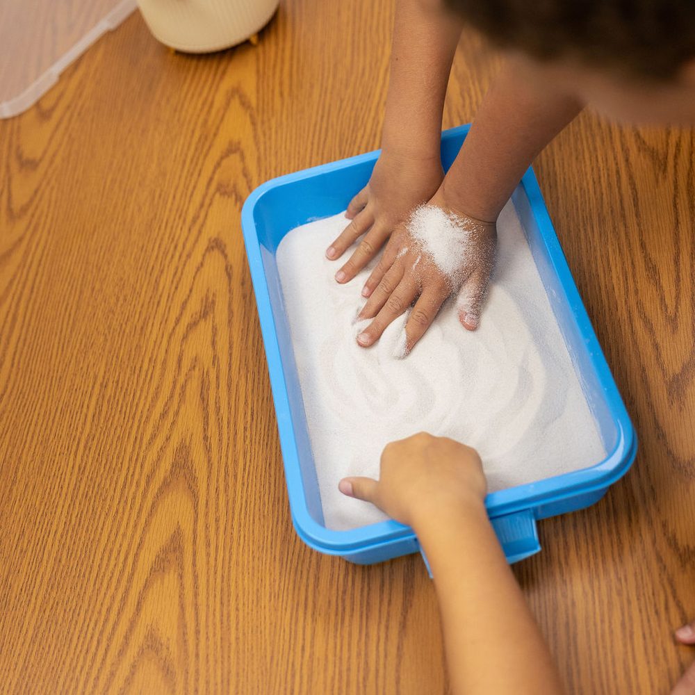 A child traces letters in a sand tray while a teacher guides the lesson, a multisensory Orton-Gillingham technique