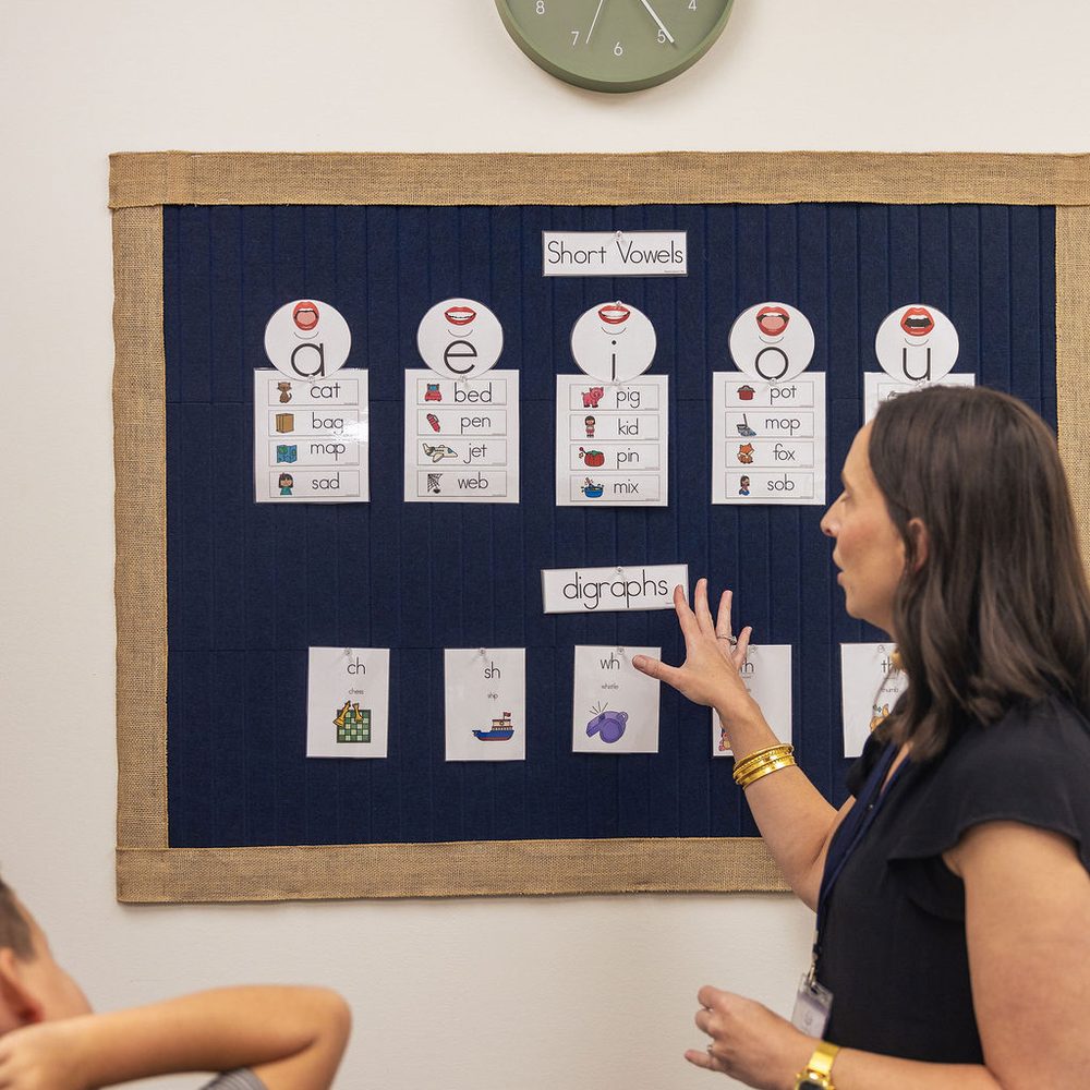 A teacher points to short-vowel and digraph cards on a classroom board during a phonics lesson